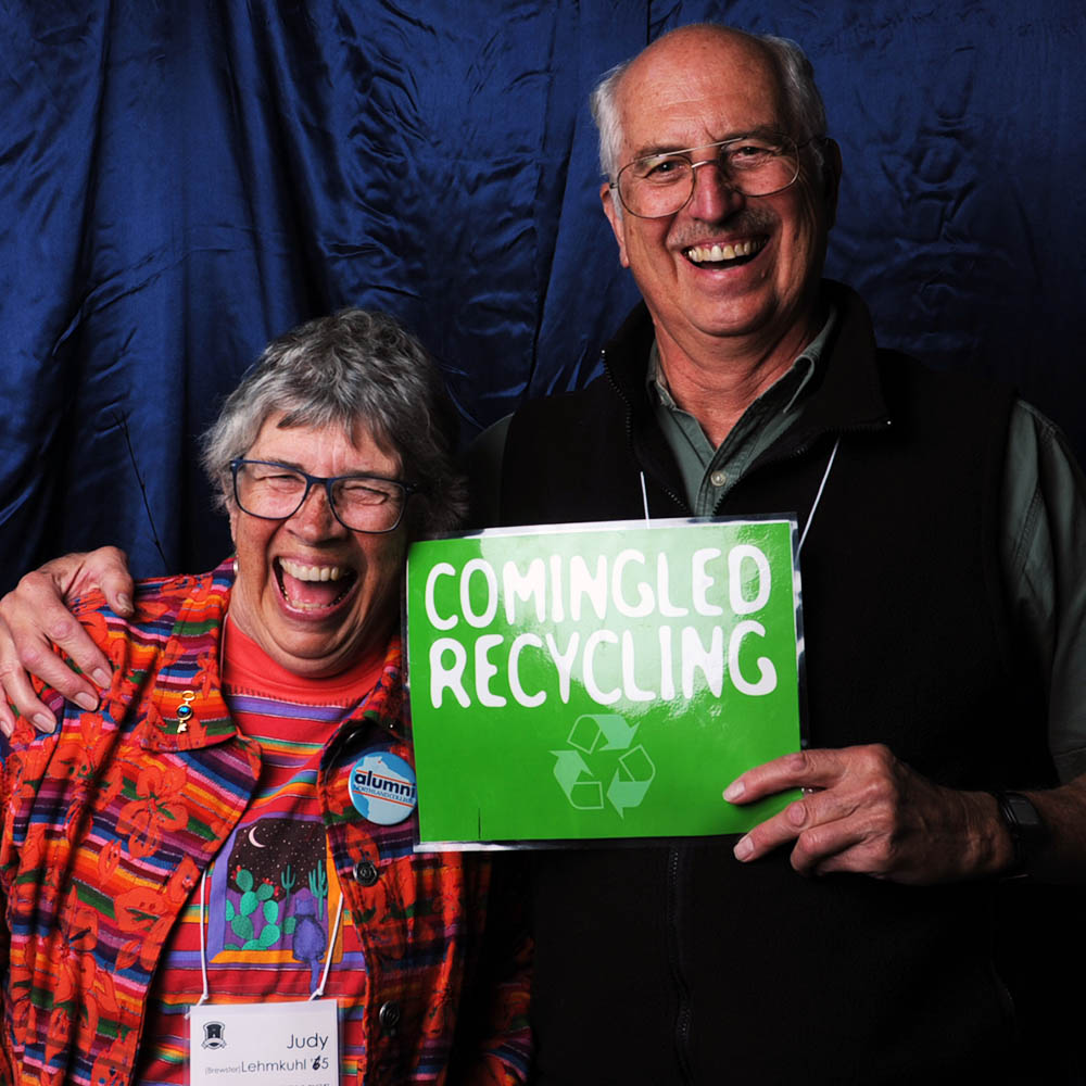 Two alumns laughing as one holds a sign that reads "Comingled Recycling"